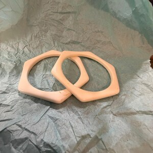 May include: Two off-white bangle bracelets with a distinct hexagonal shape. The bracelets feature a smooth, solid surface and a uniform thickness, showcasing a geometric design that creates a clean and structured appearance for the wrist accessories.