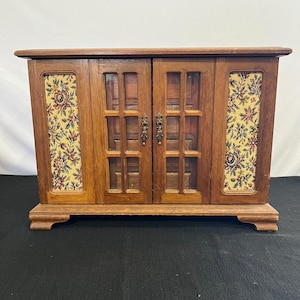 May include: A wooden jewelry cabinet with a rectangular frame and a base featuring four feet. The front has two central glass-paneled doors with metal pulls, flanked by two side panels decorated with a cream-colored fabric featuring a floral pattern.