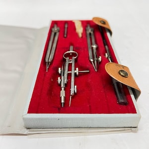 May include: A vintage drafting set in a light blue case with a red velvet interior. The set includes several silver-colored compasses and a black pen, secured by a tan leather strap with a gold-colored button. A classic tool set for technical drawing.