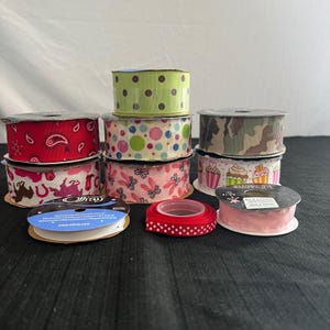 Puede incluir: Una colección de ocho carretes de cinta con varios diseños, incluyendo cachemir rojo, verde con puntos marrones, puntos multicolores, camuflaje, rosa con zapatillas de ballet, cupcakes, una cinta roja fina con puntos y una cinta rosa claro lisa.