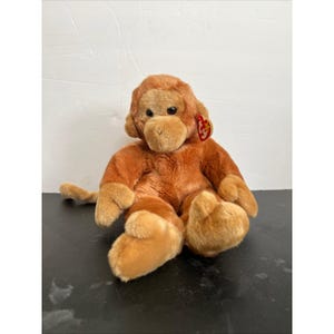 May include: A plush monkey toy with brown and tan fur, featuring a red tag. The stuffed animal has a sitting pose with its arms and legs extended. The monkey's face has a tan muzzle and dark eyes.