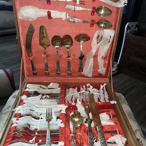 May include: An open wooden case with a red velvet interior, showcasing a complete set of antique silverware. The set includes forks, spoons, knives, and serving utensils, each with ornate silver and black handles. The silverware is wrapped in white paper.
