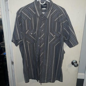 Pode incluir: Camisa de botão de manga curta com listras verticais em tons de cinza e azul. Possui dois bolsos no peito e fechos de pressão perolados. A camisa é da marca Wrangler.