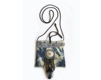 May include: A small blue and white tie-dye denim bag with a fringe and bead embellishment. The bag has a black adjustable strap and a wooden button closure.