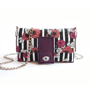 May include: A black and white striped crossbody bag with a floral print and a purple flap with a blue evil eye charm.