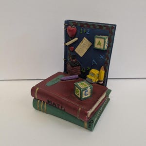 May include: A decorative figurine of stacked books with a chalkboard backdrop. The books are burgundy and green, with the word "MATH" visible. The chalkboard features an apple, numbers, and a yellow school bus. A pencil and a block with letters are also present.