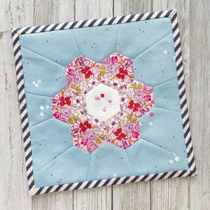 以下が含まれることがあります： 中央に花柄のデザインが施された、青と白のキルティング生地の正方形。生地は黒と白のストライプの縁取りで飾られています。