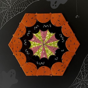 May include: A hexagonal patchwork quilt block with a central starburst design in shades of pink, green, and white. The starburst is surrounded by a ring of black fabric with a repeating pattern of small white shapes. The outer ring of the block is made of orange fabric with a black and white pattern.