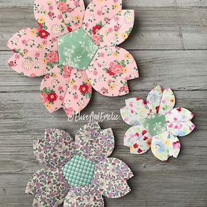 May include: Three fabric flower appliques with a green center and pink, blue, and purple floral patterns. The flowers are on a light gray wood background.