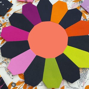 May include: A colorful fabric flower with 12 petals in shades of pink, orange, green, purple, and black. The center of the flower is a solid coral color.