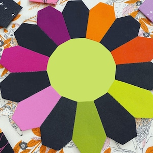 May include: A 12-pointed star-shaped fabric quilt block with alternating black, orange, green, pink, and purple fabric pieces. The center of the block is a light green circle.