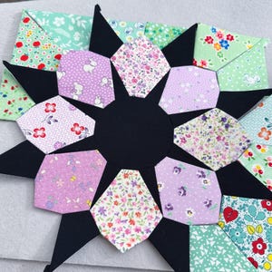 May include: A black and white quilt block with a starburst design. The block is made up of 12 smaller blocks, each with a different floral print in shades of pink, green, and white.
