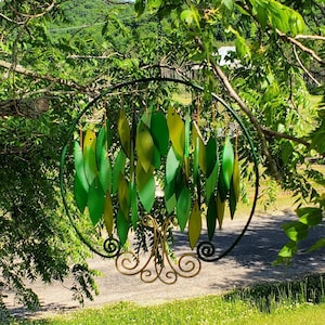 May include: A green metal wind chime with leaf-shaped pieces hanging from a circular frame. The chime is hanging from a tree branch.