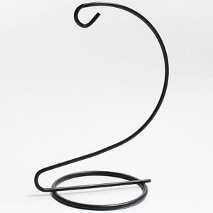 May include: A black metal decorative hanging stand with a curved hook at the top and a circular base. The stand is designed to hold items and has a minimalist, elegant design. The stand is made of metal and has a dark finish.