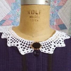 May include: A purple dress with a white crocheted collar and a black oval brooch. The dress has a button-up front and a fitted bodice.