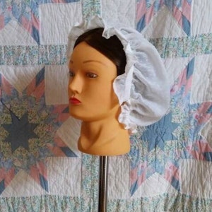 May include: A white lace bonnet with a ruffled edge. The bonnet is worn on a mannequin head.