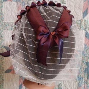 以下が含まれることがあります： 白いネットのボンネットに、ワインレッドのリボンが付いている。リボンは後ろで結ばれており、サテンの仕上げになっている。