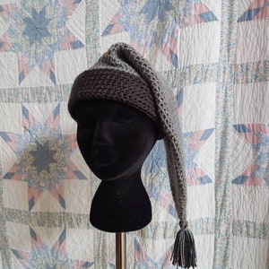 Puede incluir: Un gorro de punto gris y marrón con una larga borla gris puntiaguda. El gorro está en una cabeza de maniquí negra.