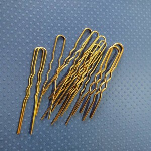 Puede incluir: Un grupo de diez horquillas doradas, tambi&eacute;n conocidas como horquillas para el cabello, est&aacute;n dispuestas en una pila sobre una superficie azul texturizada. Las horquillas son onduladas y tienen una ligera curva en el extremo.