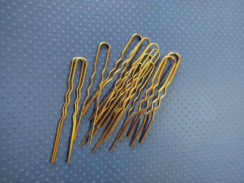 Puede incluir: Un grupo de diez horquillas doradas, tambi&eacute;n conocidas como horquillas para el cabello, est&aacute;n dispuestas en una pila sobre una superficie azul texturizada. Las horquillas son onduladas y tienen una ligera curva en el extremo.