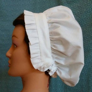 May include: A white cotton bonnet with a ruffled edge. The bonnet is gathered at the top and has a simple, classic design.