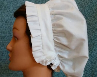 Puede incluir: Un gorro blanco de algodón con un borde de volantes. El gorro está fruncido en la parte superior y tiene un diseño simple y clásico.