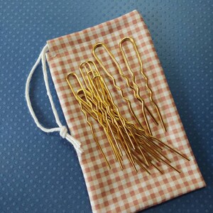 Puede incluir: Un juego de horquillas doradas en una bolsa de cord&oacute;n de gingham rosa y blanco.