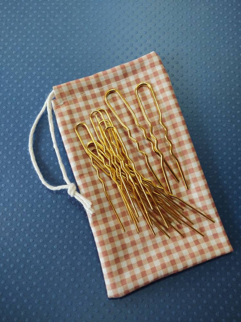 Puede incluir: Un juego de horquillas doradas en una bolsa de cord&oacute;n de gingham rosa y blanco.