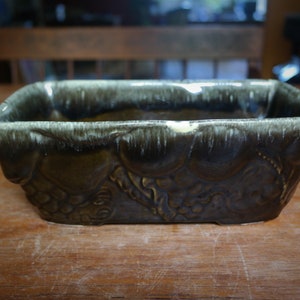 May include: A rectangular ceramic planter with a dark green glaze and a light green rim. The planter has a decorative design of leaves and vines.