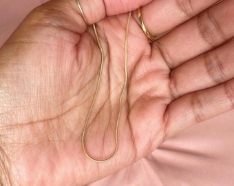 May include: A thin, gold-colored snake chain necklace with a smooth, flexible texture. The chain is draped in a gentle curve, showcasing its metallic finish and fine, interlocking link design that creates a sleek and continuous appearance.