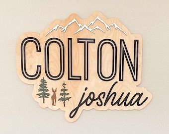 May include: A wooden sign with the names "Colton Joshua" in black lettering, with a mountain range and trees in the background. The sign is hanging on a white wall above a gray crib.