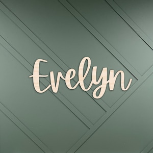 May include: A white crib with a pink and white patterned sheet, set against a green wall with a wooden sign that reads "Evelyn".