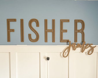 May include: A black crib with a white floral patterned sheet. The crib is against a white wainscoting wall with a brown wooden name sign that reads "FISHER" and "james" above it.