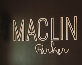 May include: A white crib with a green mattress and a brown blanket. The wall behind the crib has a wooden sign that says "Maclin Parker" in a modern font.