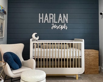 May include: A white crib with a wooden frame and a white mattress. The crib is in a nursery with a white rocking chair, a white ottoman, and a gold side table. The wall is painted a dark blue and has a white sign that says "Harlan Joseph".