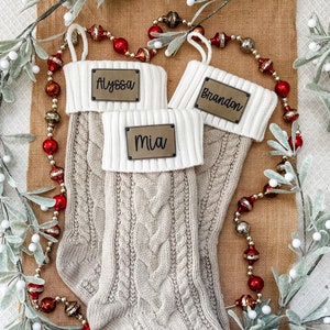May include: Three beige knitted Christmas stockings with white trim and personalized gold metal name tags. The stockings are decorated with a cable knit pattern. The names on the tags are Alyssa, Brandon, and Mia.