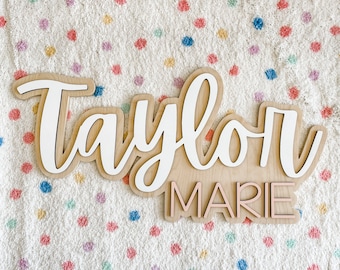 May include: Wooden sign with the names "Taylor Marie" in white and pink lettering on a light brown background.