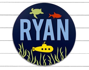 May include: A blue circular wooden sign with the name "RYAN" in light blue letters. The sign features a yellow submarine, a red fish, and a green sea turtle, all against a dark blue background.