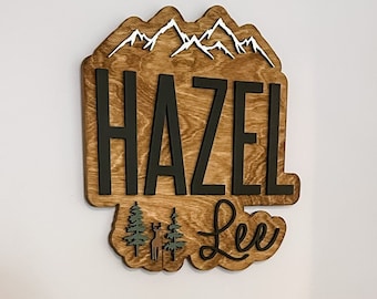 May include: A wooden wall sign featuring the name Hazel Lee in black lettering. The sign includes a white mountain range at the top and two small green pine trees with a deer silhouette at the bottom, all mounted on a natural wood backing.
