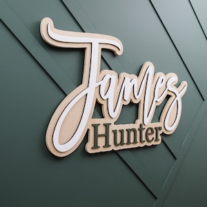 May include: A three-dimensional wall sign featuring the name James Hunter in layered wood. The word James is written in a large, white and light tan cursive script, while the word Hunter appears below in a smaller, dark green block font.