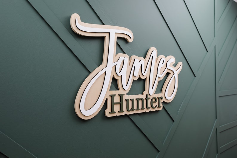 May include: A three-dimensional wall sign featuring the name James Hunter in layered wood. The word James is written in a large, white and light tan cursive script, while the word Hunter appears below in a smaller, dark green block font.