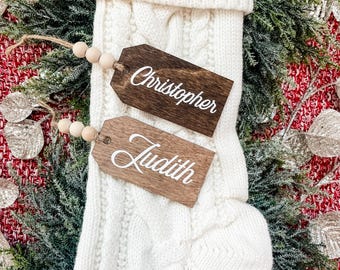 May include: Two wooden gift tags with white lettering. The tags are tied with twine and wooden beads. The tags are labeled "Christopher" and "Judith".