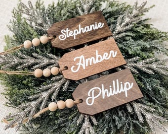 May include: Three wooden gift tags with twine and wooden beads. The tags are stained brown and have white lettering. The names on the tags are Stephanie, Amber, and Phillip.