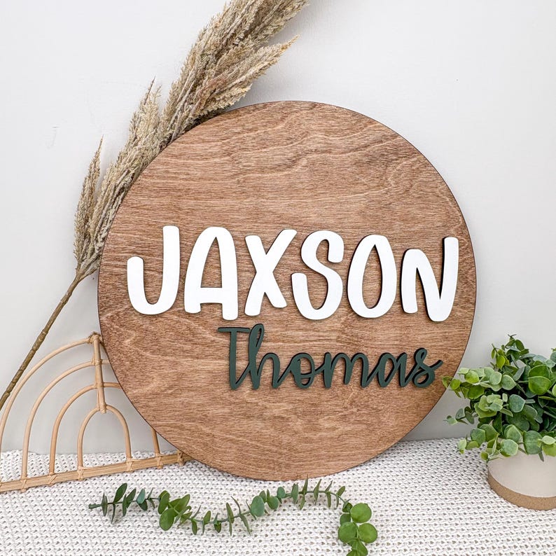 May include: A round wooden sign with the name "Jaxson" in white, raised letters above the name "Thomas" in dark green script. The sign has a natural wood grain finish and is surrounded by decorative elements.