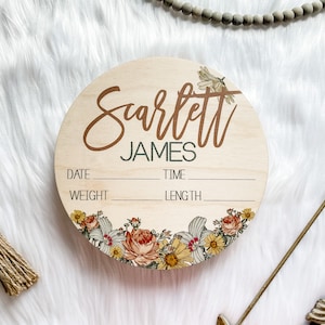 May include: A round wooden baby announcement sign with the name "Scarlett James" in brown script. The sign includes spaces for date, time, weight, and length. Floral designs in yellow, orange, and white adorn the bottom edge.