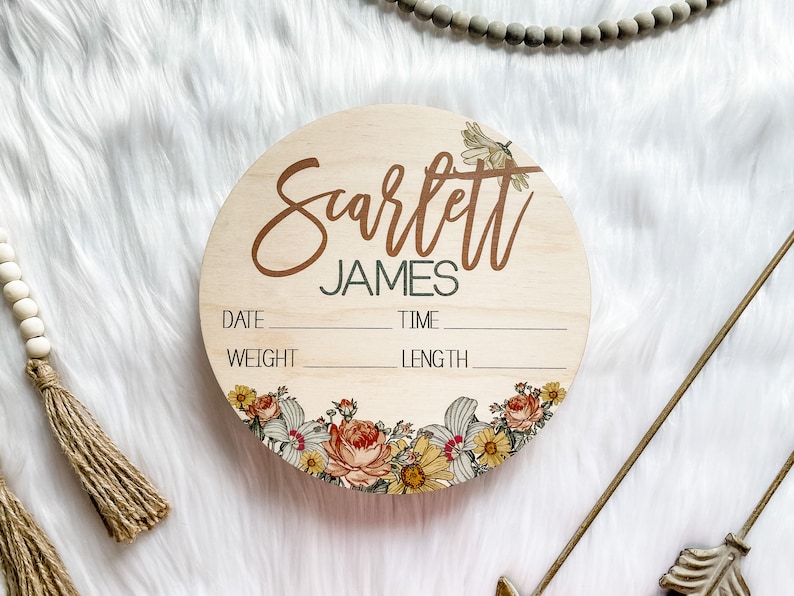 May include: A round wooden baby announcement sign with the name "Scarlett James" in brown script. The sign includes spaces for date, time, weight, and length. Floral designs in yellow, orange, and white adorn the bottom edge.