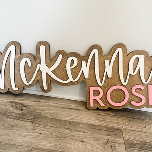 May include: Wooden sign with the name "Mckenna Rose" in white and pink letters.