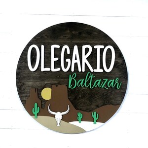 K&ouml;nnte beinhalten: Ein rundes Holzschild mit braunem Hintergrund und einem in Wei&szlig; lackierten Namen "OLEGARIO Baltazar". Das Schild zeigt eine W&uuml;stenlandschaft mit einem braunen Berg, einer gelben Sonne, einem wei&szlig;en Kuhsch&auml;del und gr&uuml;nen Kakteen.