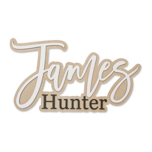 May include: A custom wall sign featuring the name James in a large, white, cursive script layered over a light wood-toned backing. Below the name James, the word Hunter is displayed in a smaller, bold, dark grey sans-serif font.
