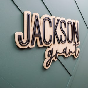 May include: A custom wall sign featuring the name JACKSON in bold, black, sans-serif capital letters layered over a light wood backing, with the name grant in a black, cursive script font layered below, creating a three-dimensional effect.